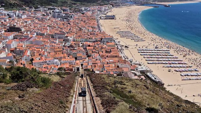 Two Nazar&eacute; funicular cars cross on the hillside, with one ascending while the other simultaneously travels in the opposite, descending direction toward the town and beach in Portugal.