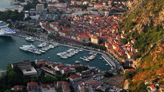aerial drift across a mountain slope in low evening sun with warm light on the red roofs and stone walls of kotor old town and dark calm water in the marina, montenegro