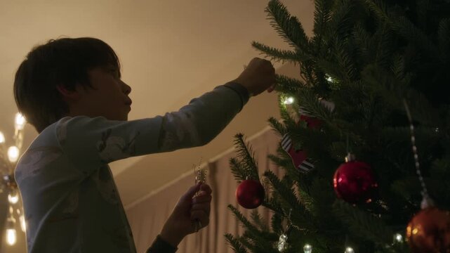 Young boy hanging up reusable tinsel on indoor christmas tree at night preparing for holiday