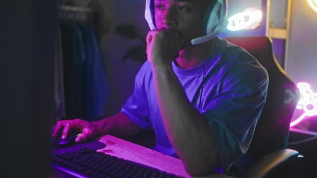 Man with white headset using mouse at desktop computer in studio, neon bitcoin and controller signs, shelving and plant behind him; concentration competition.