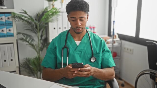 Man doctor in green scrubs seated at desk holding smartphone with stethoscope visible in building; calm focused professionalism.