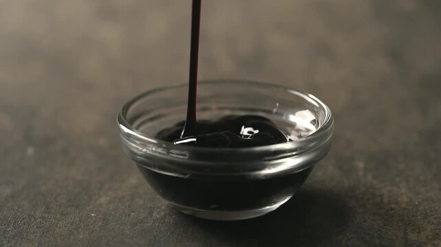 Thick dark sauce pouring into a small clear glass bowl on a textured stone table. Static close-up studio shot with soft lighting, dark moody background, and copy space.