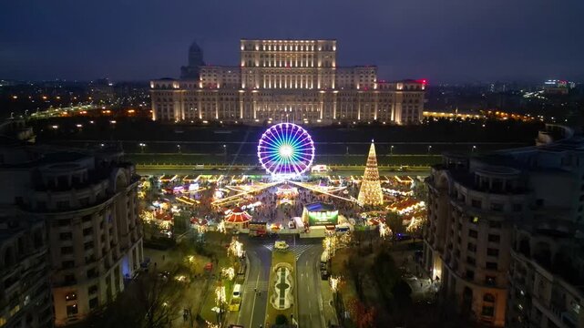 Aerial drone footage of the festive Bucharest Christmas Market illuminated at night in Constitution Square (Piata Constitutiei), featuring the glowing Ferris wheel, decorated Christmas tree.