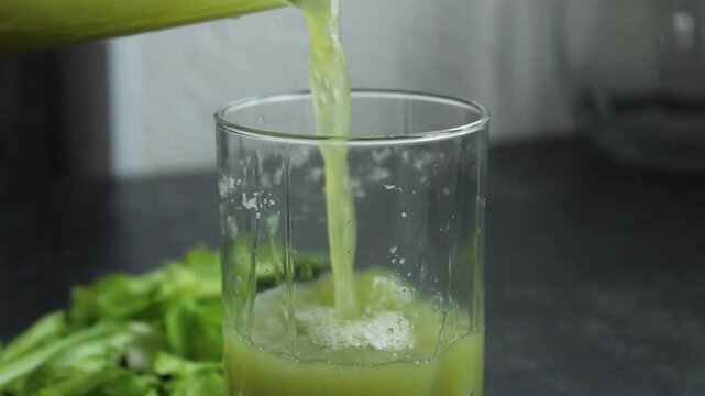 Fresh green celery juice is poured into a clean glass from a pitcher, creating a healthy and refreshing drink.