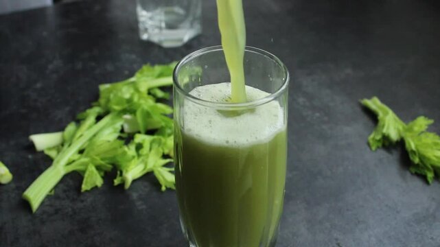 close-up shot of fresh celery juice being poured into a glass, with celery stalks scattered on a dark surface.