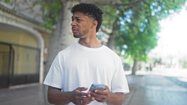 Man holding smartphone scrolling with thumbs and frowning while looking down on street near building arch; concerned solitude.