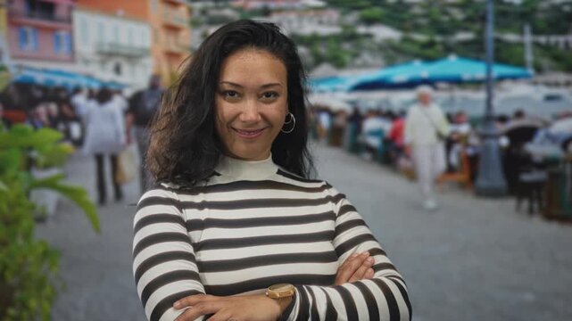 Woman with arms crossed smiling and showing teeth on a street terrace cafe with umbrellas, outdoor tables and diners visible; confidence optimism.