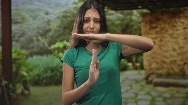 Young hispanic woman holding hands in time out gesture beside stone building in forest; frustration pause resilience.