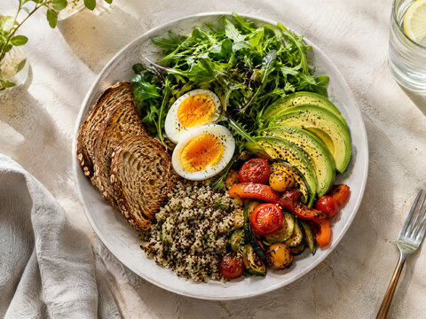 Healthy breakfast bowl with soft boiled eggs, fresh avocado slices and quinoa. Nutritious morning meal with toasted sourdough bread and roasted vegetables. Vegetarian brunch plate.