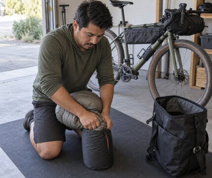 Cyclist compressing a lightweight sleeping quilt into a stuff sack on a garage mat before an overnight bikepacking trip with an open luggage bag and a bicycle carrying compact frame storage