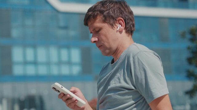 Man with wireless earbuds checking smartphone outdoors while listening to podcast or audiobook, concept of audio learning, mobile education and digital lifestyle.