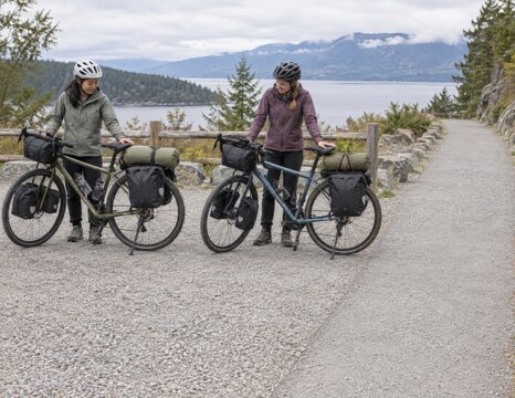 Two helmeted bikepackers park loaded touring bicycles in parallel within a broad gravel turnout while checking their spacing and leaving the adjacent walking path clear during an overnight trip