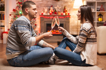 Naklejka premium Man and woman clink champagne flutes beside a Christmas tree and fireplace, sharing a festive holiday toast and intimate celebration at home.