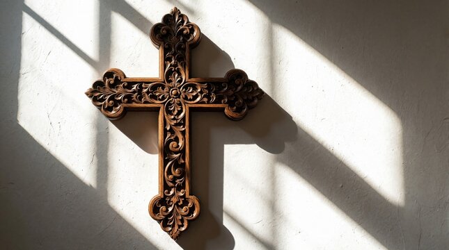 Ornate wooden cross hangs on textured wall. Sunlight creates patterns and shadows on wall behind carved religious symbol. Interior home decor accent.