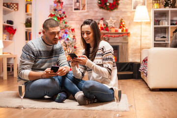Obraz premium Couple sits close together on the floor using separate smartphones beside a decorated tree and fireplace during relaxed Christmas downtime at home.