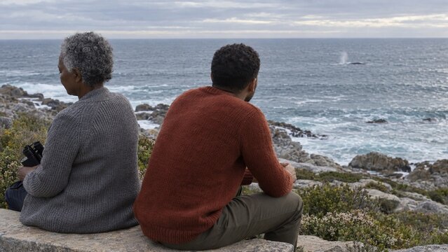 two people sit on a stone ledge, one person holds binoculars while looking at the ocean, waves crash against rocks under a cloudy sky