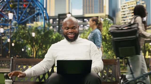 Businessman drinks coffee from a cup while using a laptop on a park bench, combining remote work with a break in an urban public park and a flexible mobile office lifestyle outdoors.