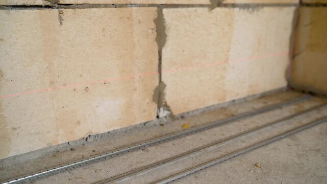 Camera moves along metal floor guides set for a screed. A red laser level projects a precise line on an unfinished aerated concrete block wall during renovation or construction