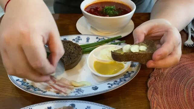 Close-up of a person's hands making a traditional Slavic sandwich, spreading garlic and mustard on a piece of dark rye bread, and putting thinly sliced pork fat salo on top, with a plate of fresh 