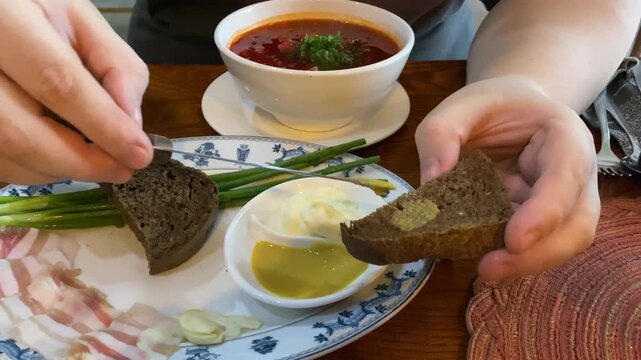 Close-up of a person's hands using a knife to spread spicy yellow mustard on a slice of dark rye bread, preparing a traditional appetizer to accompany a hot bowl of Ukrainian borscht soup, salo pork f