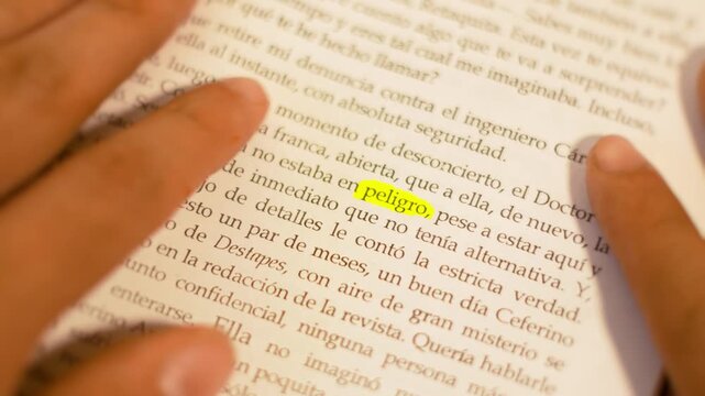 Macro close-up of a hand flipping book pages until reaching the yellow highlighted word 'peligro' under dark lighting.