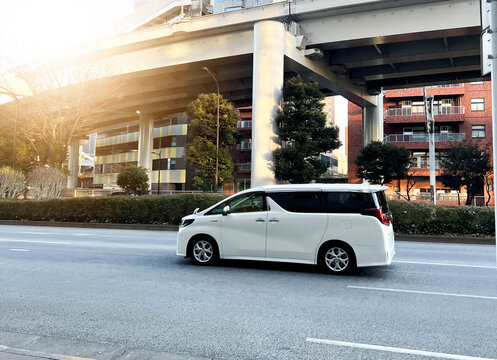 A white Toyota Alphard luxury minivan drives along a street in Japan under an elevated highway overpass during golden hour.