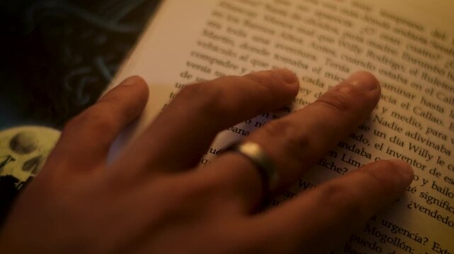 Macro detail of a hand wearing a ring gently touching a classic book page under warm low-key lighting.