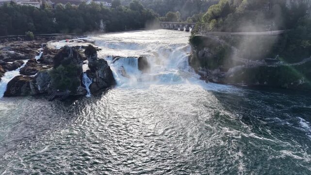 Aerial view of the Rhine Falls in Switzerland with powerful cascading water, rocky formations, rising mist, viewing platforms and historic railway bridge near Schloss Laufen.
