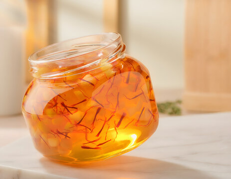 Golden saffron-infused honey in a clear glass jar, a gourmet culinary ingredient; vibrant orange saffron threads steeped in viscous, amber-colored honey