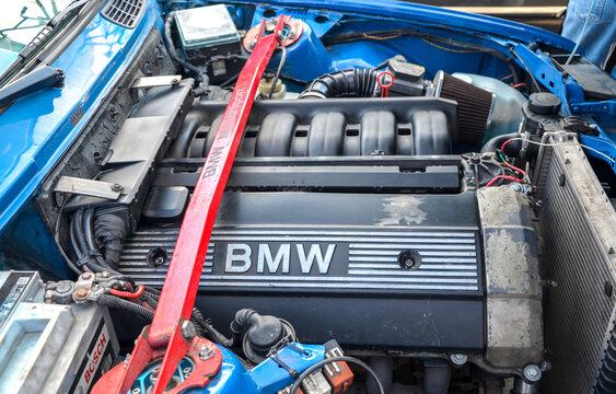 Close up of a tuned BMW inline-six engine bay with a red strut bar, custom intake filter, and Bosch battery inside a blue sports car chassis