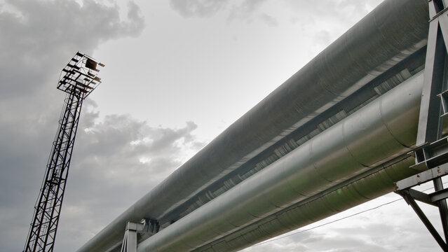 The photo shows a pipeline against a gray sky, and a tower with floodlights in the background