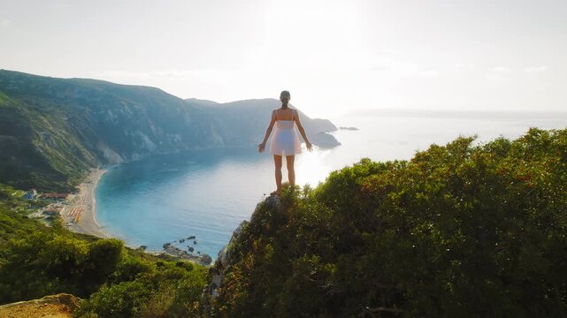 Woman in white dress admires sunrise over Ionian Sea from high cliff in Kefalonia