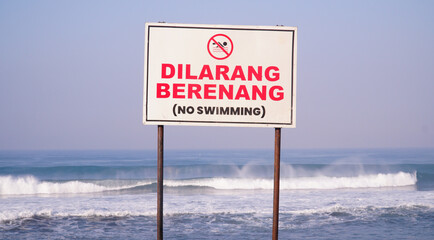 An Indonesian and English "Dilarang Berenang - No Swimming" warning sign stands on a black sand beach with ocean waves and blue sky in the background © tarasari