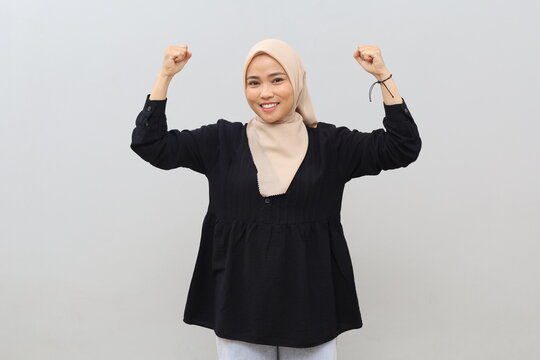 A portrait of an attractive and confident young Asian Muslim woman wearing a headscarf, flexing her biceps to show her strength, with a stylish smile and a bold attitude.