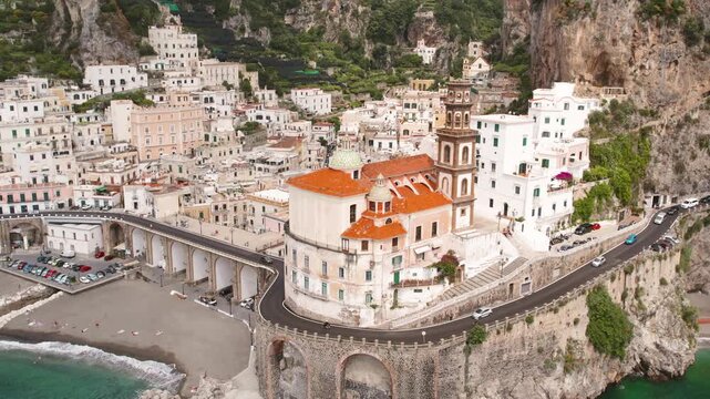 Aerial view of Atrani city with a prominent church, intricate architecture and buildings by the sea and mountains in the background.