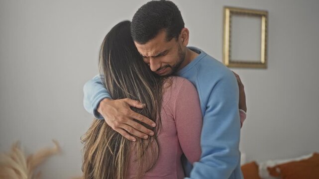 Man hugs woman, hand on back and palm on shoulder inside building living room with framed picture, couch and pampas grass; comfort.