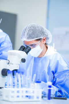 A female microbiologist in a white coat, protective eyewear and a sterile hairnet settling alone at the stereo microscope inside a university biotechnology laboratory.