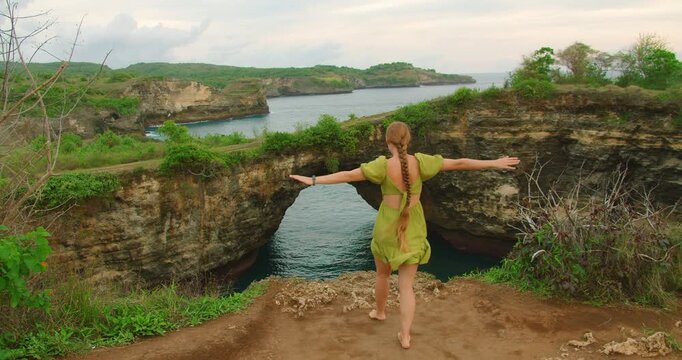 Woman tourist walking with arms outstretched on cliff edge overlooking Broken Beach arch Nusa Penida Bali
