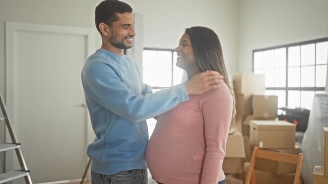 Man smiling hugging pregnant woman, hand on belly and kissing cheek amid moving boxes in building; anticipation joy.