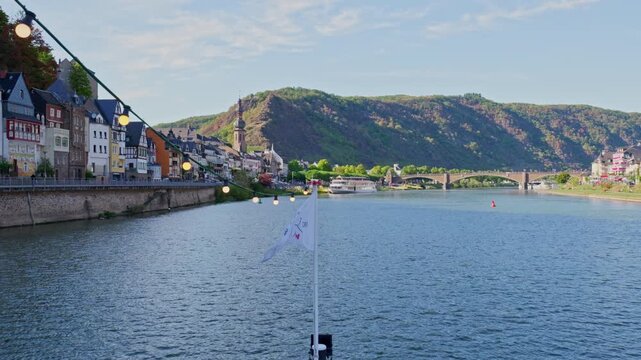 Cinematic wide video of the scenic Moselle river flowing through the historic town of Cochem in Germany, featuring traditional waterfront architecture, a stone bridge in the background, a white flag, 
