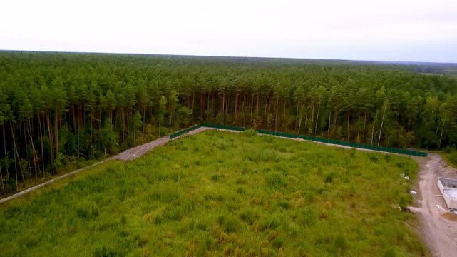 Aerial view of a green field bordering a pine forest with a metal fence enclosure
