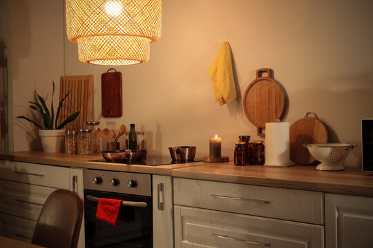 Interior of modern kitchen with glowing lamp, oven and utensils on white counter at evening