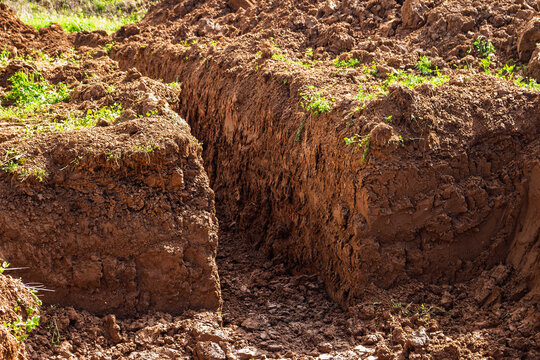 Deep excavation trench dug into the earth for a military bunker, showing layers of brown clay soil and rugged ground texture created by heavy machinery works in a field setting