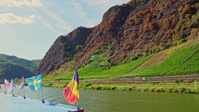 Scenic mountain cliff and vineyards along a river with flags on a boat during a summer cruise
