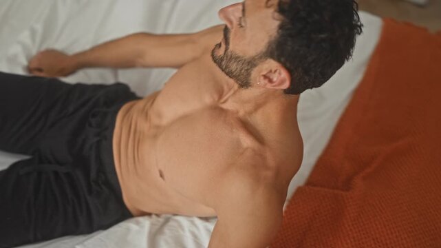Middle aged man, shirtless, with bare back turning head while sitting on bed in a bedroom of a residential building; quiet introspection.