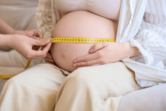 Young pregnant woman with doula measuring belly on sofa at home, closeup