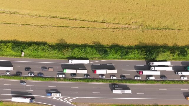 Aerial perspective of a congested highway featuring trucks and cars traveling in both directions, bordered by a field and lush greenery