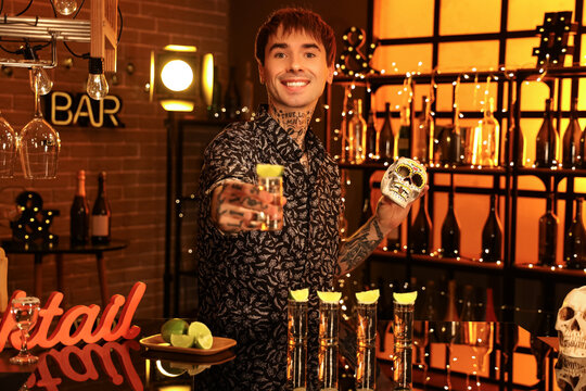 Young tattooed man with tequila shot and painted skull in bar at night