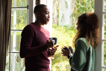 Naklejka premium Diverse couple standing by French door, holding mugs, talking with smartwatch visible