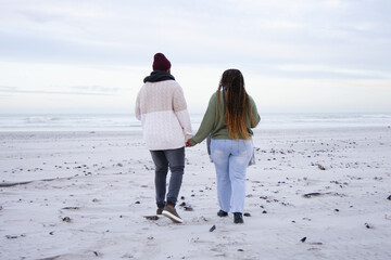 Obraz premium African American couple holding hands walking to sea on beach in knits leaving footprints, pebbles